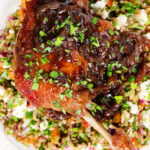 Overhead close-up baked pomegranate molasses duck leg with onions, served with a feta freekeh salad.