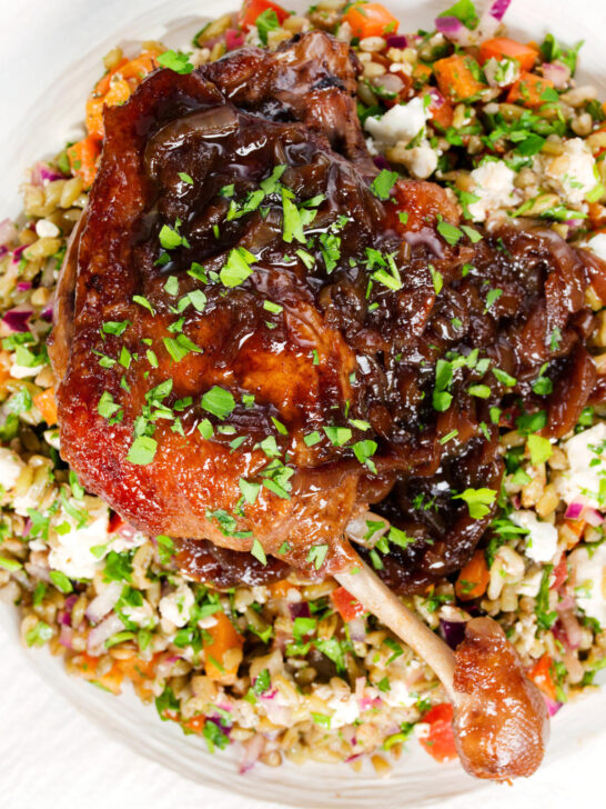 Overhead close-up baked pomegranate molasses duck leg with onions, served with a feta freekeh salad.