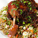 Close-up baked pomegranate molasses duck leg with onions, served with a feta freekeh salad.