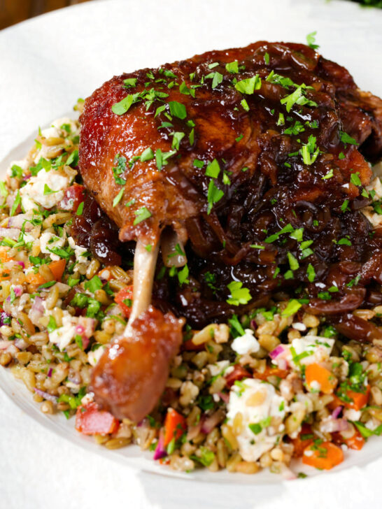Close-up baked pomegranate molasses duck leg with onions, served with a feta freekeh salad.