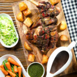 Overhead slow roasted lamb shoulder with roast potatoes, veggies, mint sauce and gravy served on a board.