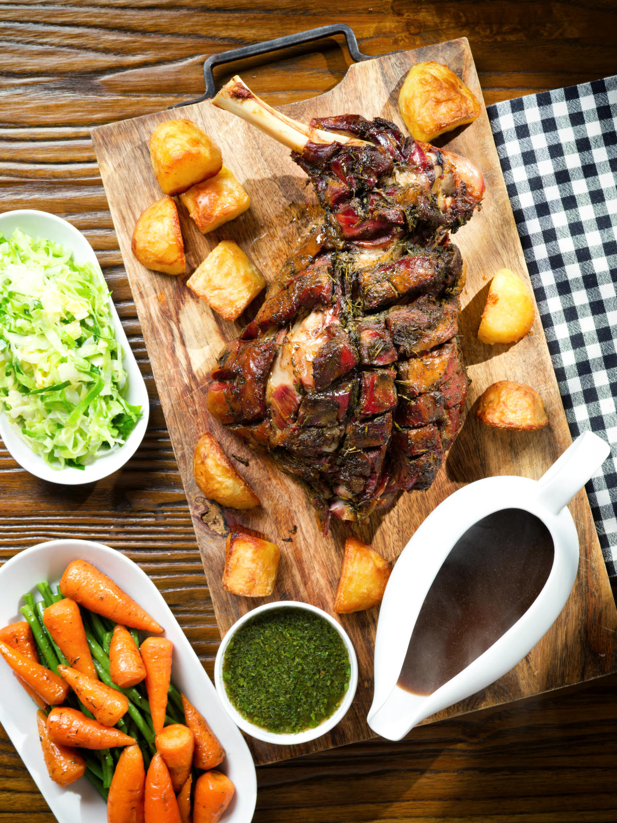 Overhead slow roasted lamb shoulder with roast potatoes, veggies, mint sauce and gravy served on a board.