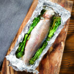 Overhead whole baked fish (rainbow trout) cooked in foil with asparagus and tenderstem broccoli.