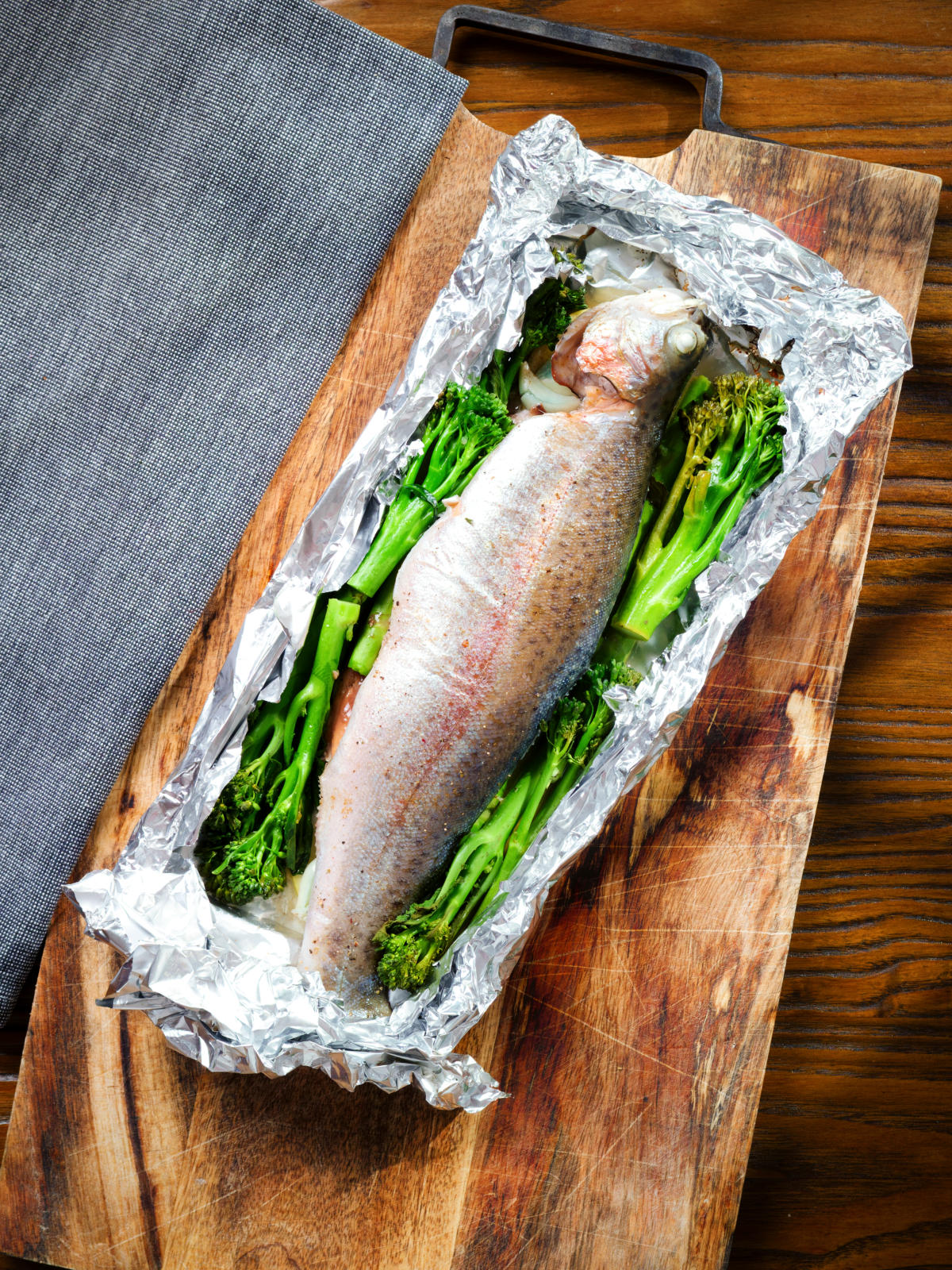 Overhead whole baked fish (rainbow trout) cooked in foil with asparagus and tenderstem broccoli.