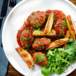 Overhead soutzoukakia (Greek beef meatballs) in tomato sauce with salad and potato wedges.