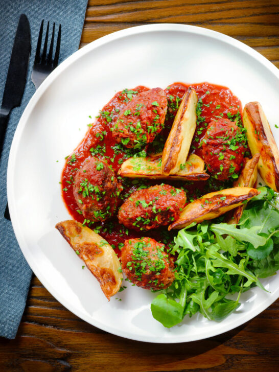Overhead soutzoukakia (Greek beef meatballs) in tomato sauce with salad and potato wedges.