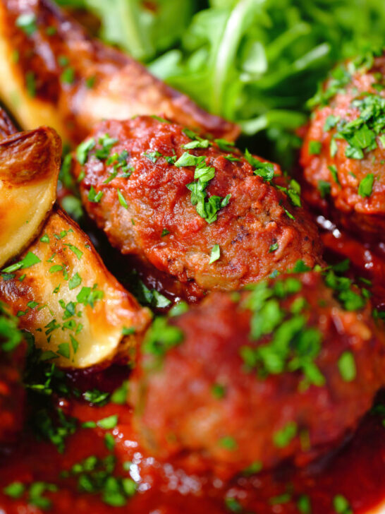Close-up Soutzoukakia (Greek beef meatballs) in tomato sauce with baked potato wedges.