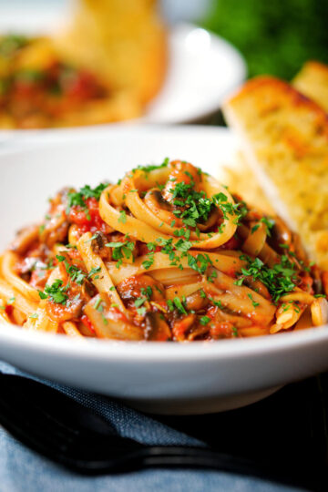 Close-up tomato and mushroom pasta garnished with parsley served with garlic bread.