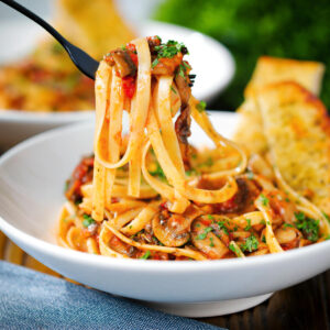 Tomato and mushroom pasta (fettucine) on a fork served with garlic bread.