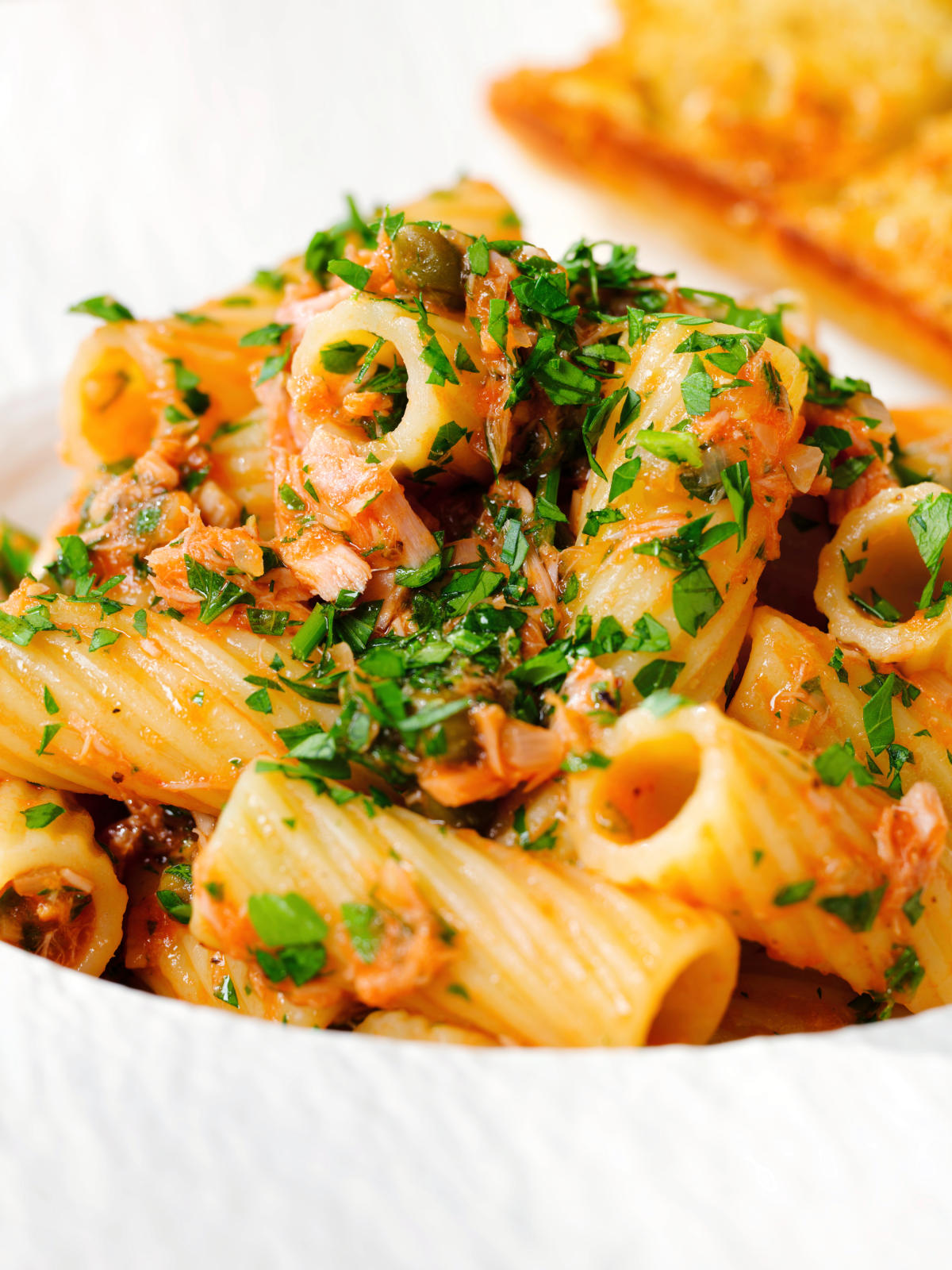 Close-up tinned tuna tomato pasta with capers, served with garlic bread.